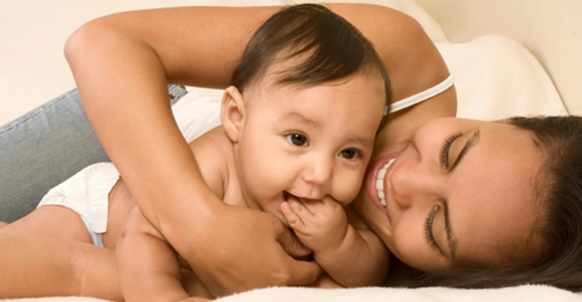 Smiling mother lying on a bed cuddling her baby, who is looking forward with a hand in their mouth.
