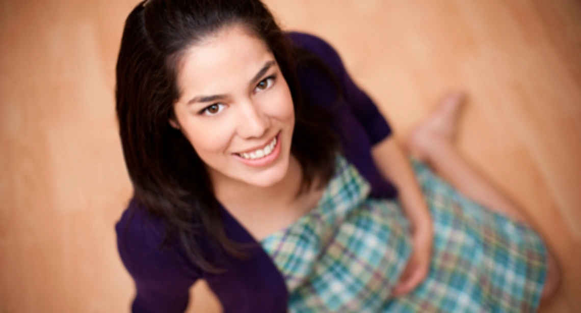 Smiling pregnant woman sitting on the floor, wearing a green and blue plaid dress with a purple sweater.