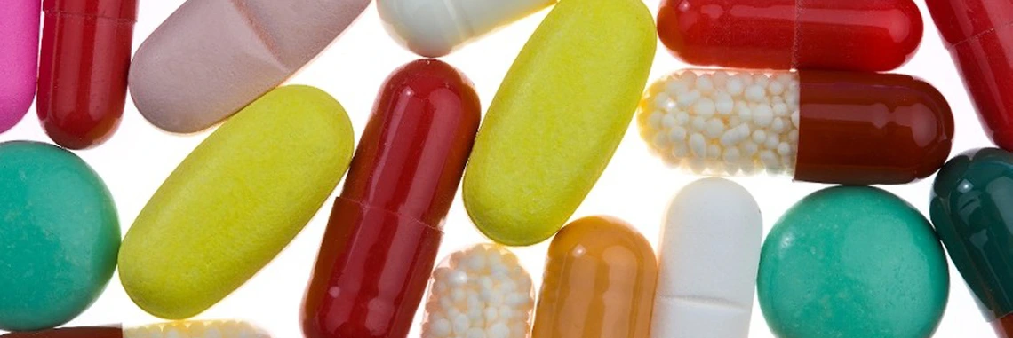 Colorful assortment of pills and capsules, including round tablets and oblong gel caps, arranged closely together on a light background.