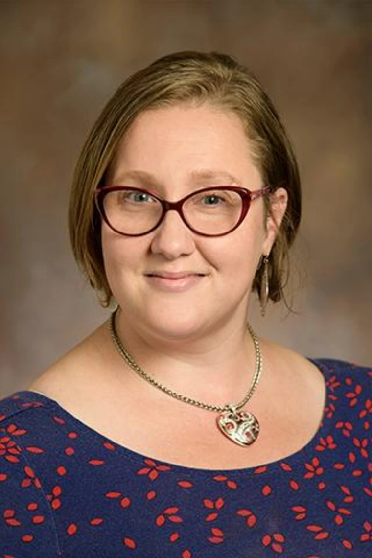 Person with short hair and glasses wearing a navy dress with red pattern and a silver heart necklace.