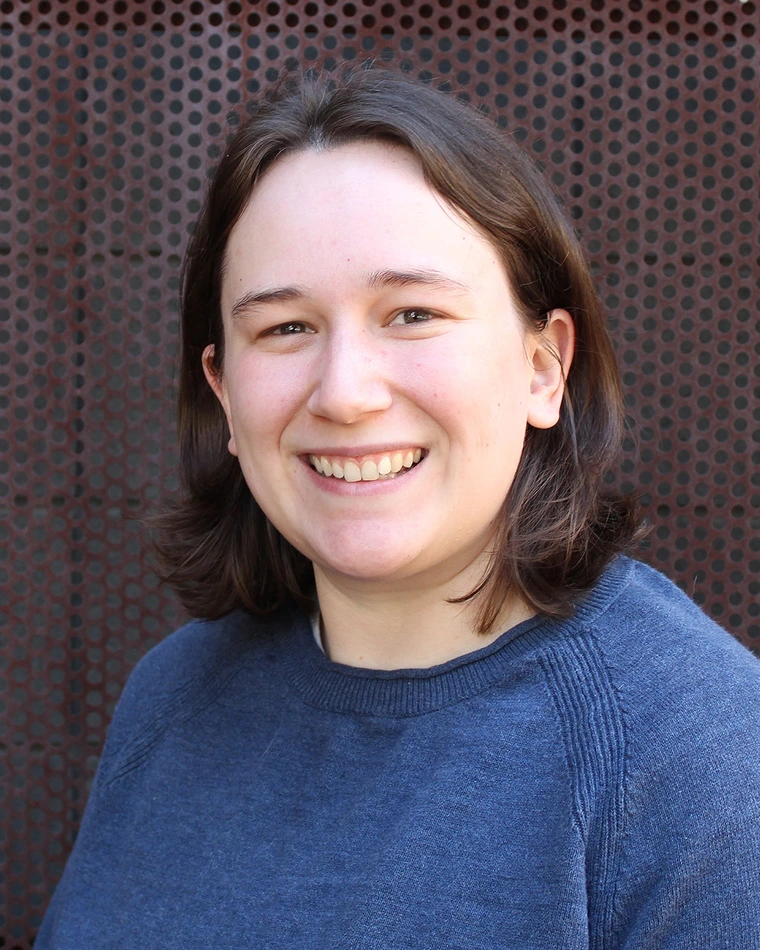 Person with short brown hair wearing a blue sweater smiling in front of a perforated metal background.