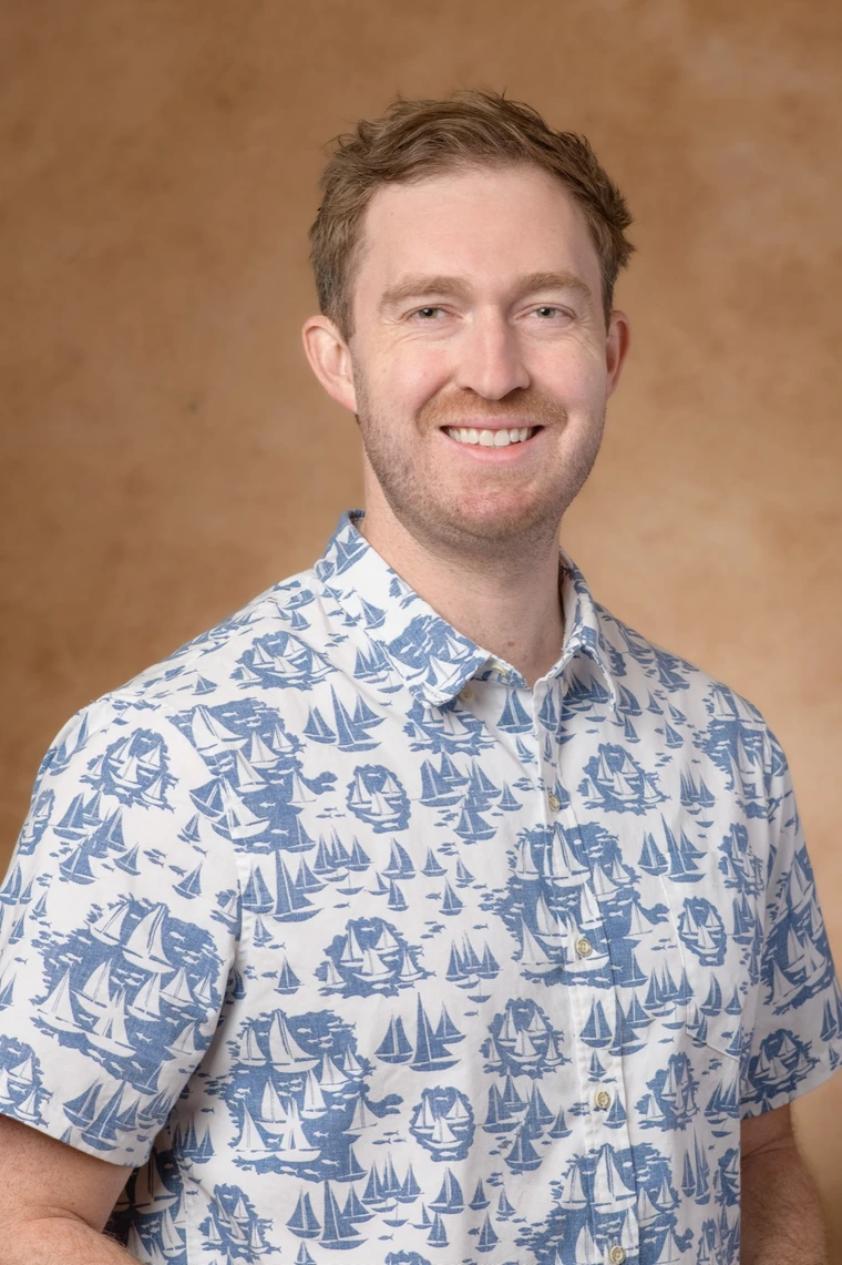 Person with short reddish-brown hair wearing a white shirt with a blue sailboat print, smiling in front of a tan background.