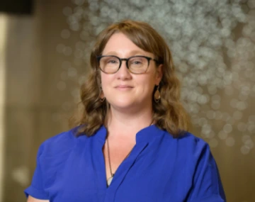 Woman wearing glasses and a blue blouse standing in front of a softly blurred background with circular light reflections.