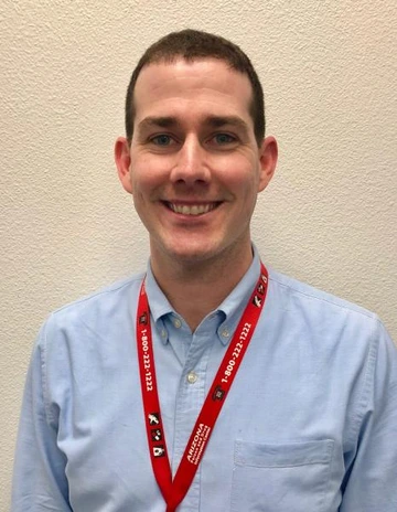 A smiling man wearing a light blue shirt and a red Arizona Poison and Drug Information Center lanyard standing against a neutral wall.
