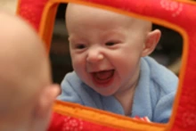 Laughing baby in a blue outfit looking at their reflection in a mirror.