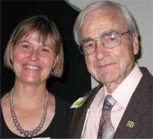 Two people smiling for a photo together, one wearing a necklace and the other wearing glasses, a suit, and a tie.